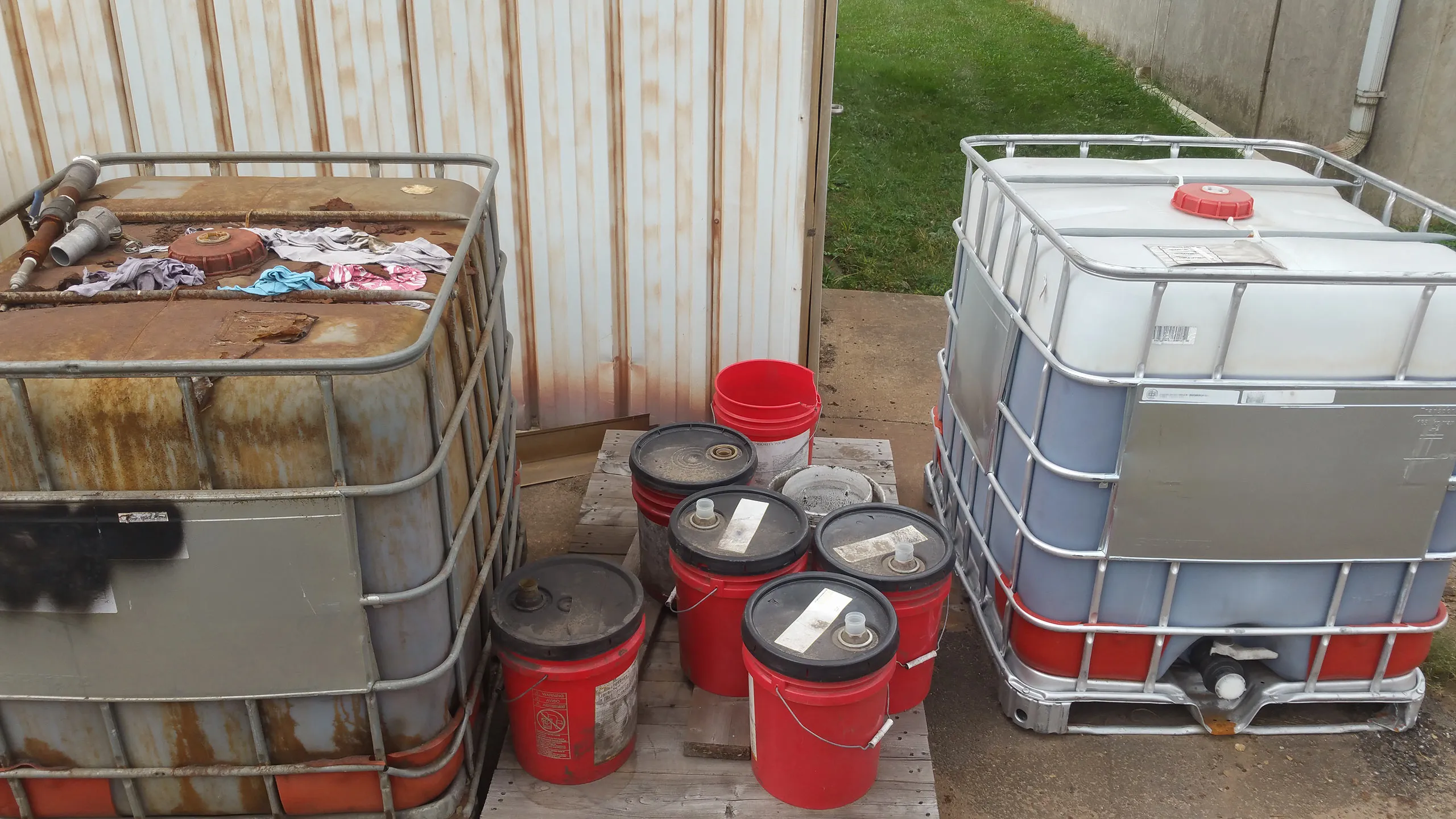 Two intermediate bulk containers with sealed five-gallon pails on a pallet between them, staged outdoors for collection.