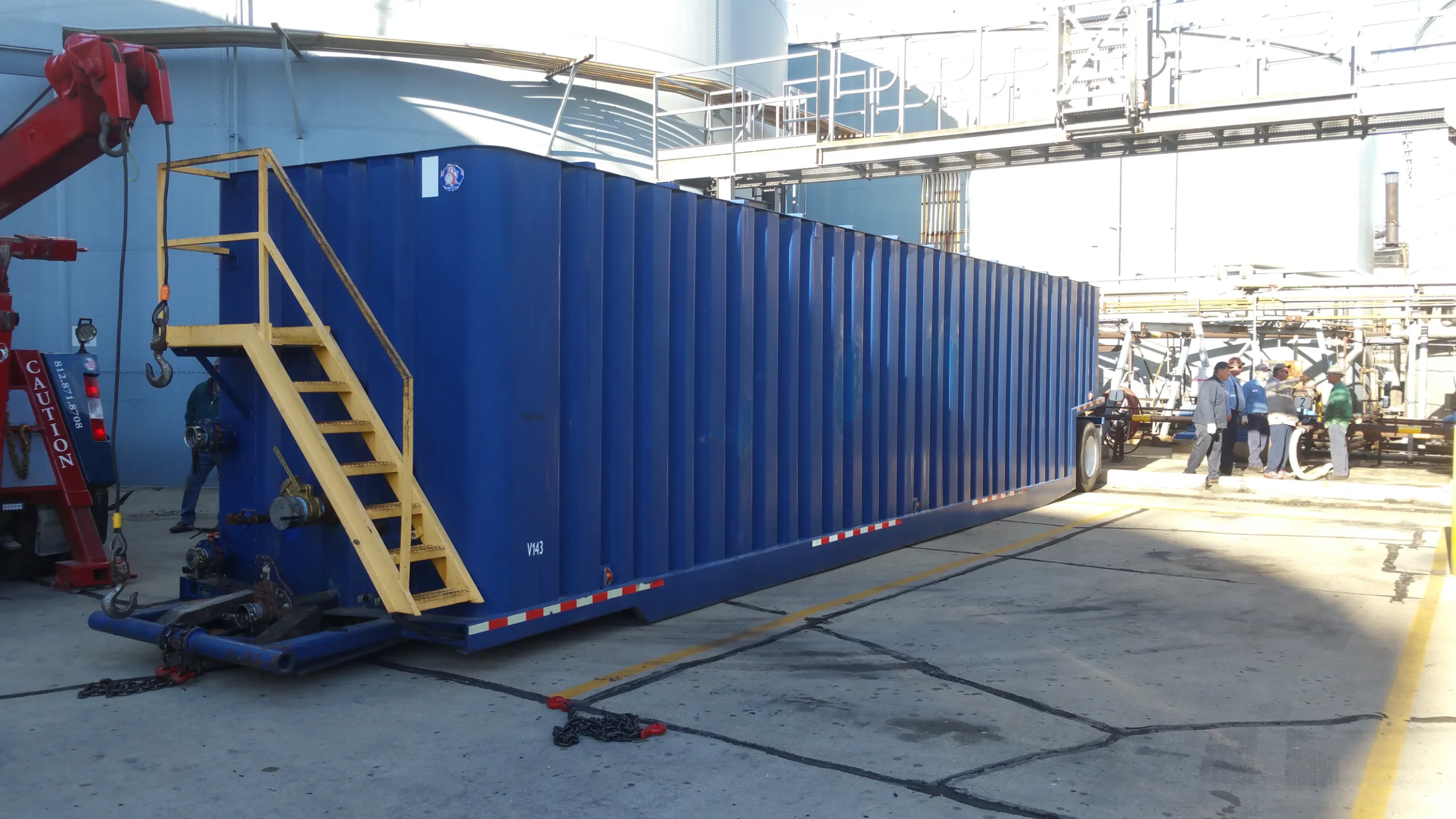 A blue roll-off tank container with access stairs being positioned by a hook truck at an industrial plant.