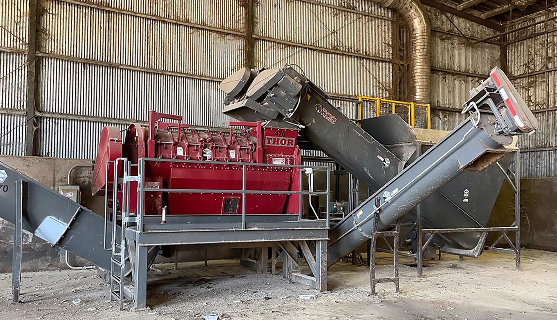 An industrial shredder line on a steel platform, fed and discharged by inclined conveyors inside the processing building.