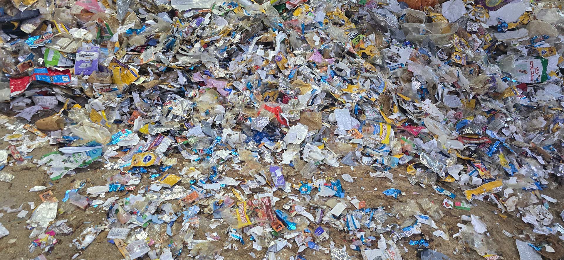 A wide spread of mixed flexible packaging waste — foil pouches, film wrappers and label scrap — covering a concrete floor before sorting.