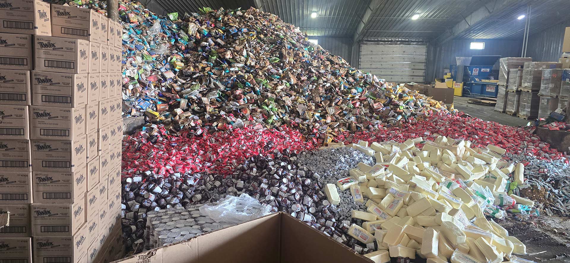 Warehouse floor piled with out-of-spec packaged food, separated into distinct streams beside stacked cases and a baler.